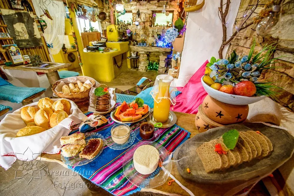 Mesa do café da manhã com pães, broas, mamão, suco, queijo e geleia sobre toalha colorida
